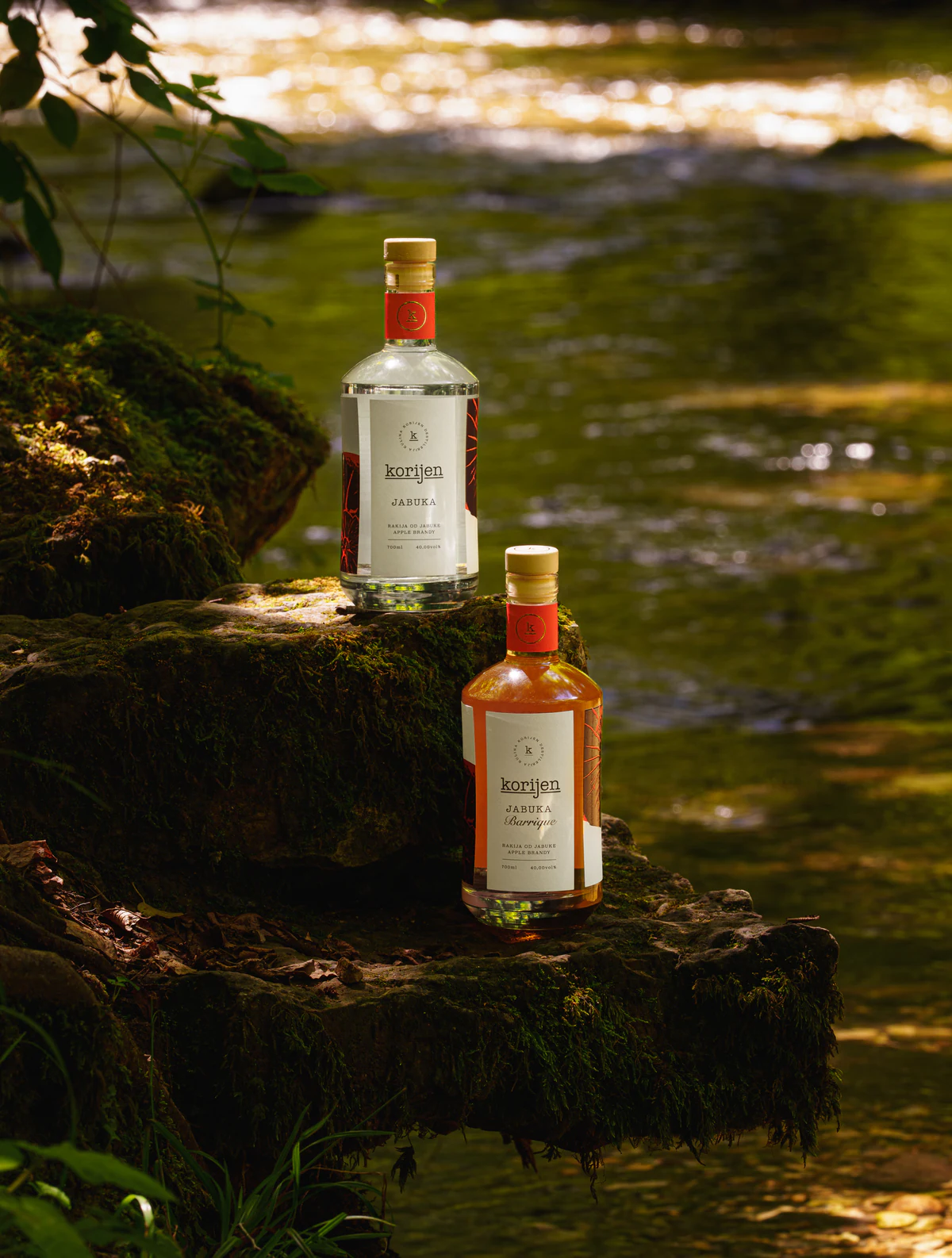 Clear and barrique Korijen bottles arranged beside running water in a woodland setting.