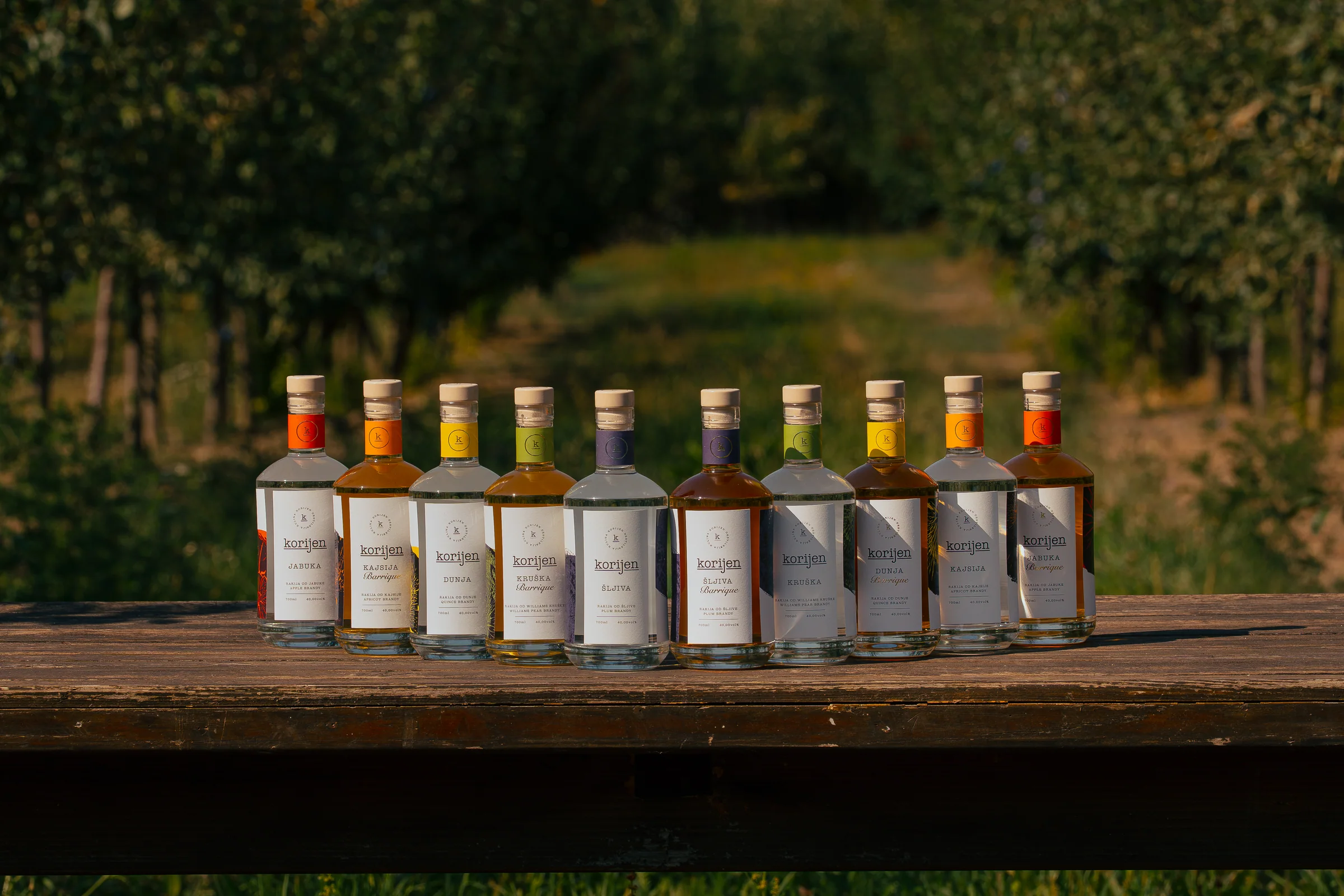 All ten Korijen rakija bottles arranged on a wooden table in the orchard.